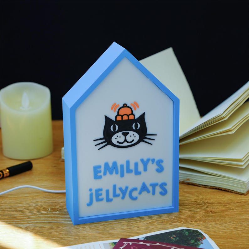May include: A light blue house-shaped night light with a black cat illustration wearing a crown. The text "EMILLY'S JELLYCATS" is displayed in blue. The night light is on a wooden surface with a candle and book in the background.