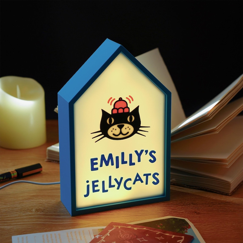 May include: A blue house-shaped night light with the text "EMILLY'S JELLYCATS" and a black cat graphic. The light is on, illuminating the text and graphic. A candle and books are in the background.