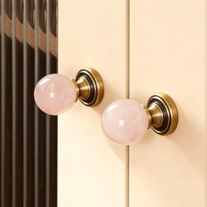 May include: Close-up of a cream-colored cabinet door with two pink quartz crystal knobs. The knobs have a brass base and are mounted on a cabinet with a ribbed glass panel.