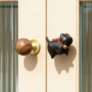 May include: A wooden doorknob with a brass base and a black cat-shaped door pull. The cat has a red collar and a small gold bell. The door is a light cream color with a ribbed glass panel.