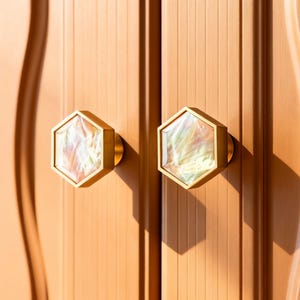 May include: Close-up of two cabinet door knobs. Each knob has a hexagonal shape with a gold-colored frame and a pearlescent, iridescent center. The knobs are mounted on a light brown cabinet door with vertical grooves.