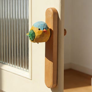 May include: A wooden door handle with a carved bird design. The bird is painted in shades of yellow, blue, green, and orange. The handle is attached to a light-colored wooden door with a glass panel.