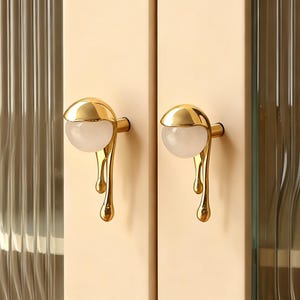 Könnte beinhalten: Zwei Schranktürgriffe. Jeder Griff hat eine goldfarbene, abgerundete Oberseite mit einem weißen, kugelförmigen Zentrum. Unterhalb der Oberseite tropft das Goldmetall in einer stilisierten, flüssigen Form herunter. Die Griffe sind an einer hellbeigen Schranktür befestigt.