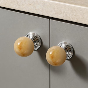 Könnte beinhalten: Nahaufnahme von zwei Schrankknöpfen. Jeder Knopf hat eine hellbraune, marmorähnliche Kugel auf einem silbernen, geriffelten Sockel. Die Knöpfe sind an einer grauen Schranktür befestigt. Im Hintergrund ist eine beige Arbeitsplatte zu sehen.