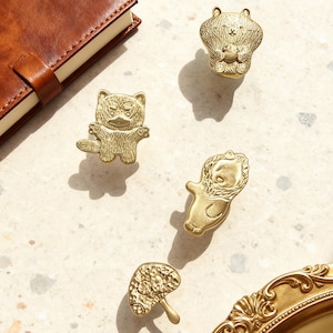 Könnte beinhalten: Eine Sammlung von Schrankknöpfen in Form von Tieren aus Messing, darunter ein Bär, ein Waschbär, ein Löwe und ein Pilz. Die Knöpfe sind auf einer hellen Oberfläche angeordnet, in der Nähe eines ledergebundenen Notizbuchs und einer dekorativen goldenen Schale.