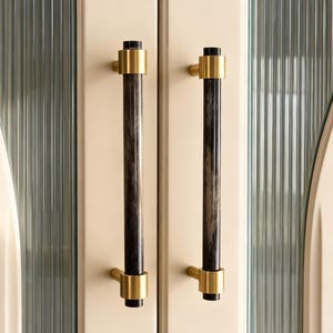 Puede incluir: Primer plano de dos tiradores de puerta de armario. Cada tirador tiene un centro cilíndrico oscuro con detalles dorados en la parte superior e inferior. Los tiradores están montados en una puerta de armario color crema con un panel de vidrio texturizado.