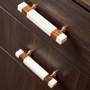 Peut inclure: Deux poignées de meubles rectangulaires. Les poignées présentent une section centrale blanche, flanquée de bois foncé et d'embouts blancs. Elles sont montées sur une armoire brun foncé avec une quincaillerie dorée.