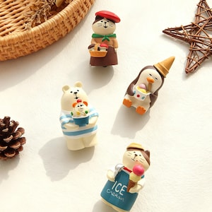 May include: A collection of whimsical figurines, including a cat in a beret, a penguin with an ice cream cone, and a bear holding a small bowl. The figurines are in various poses and colors, with a light background.