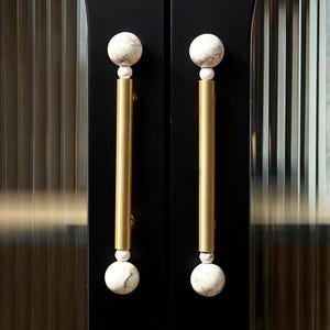 May include: Close-up of two cabinet door handles. Each handle features a gold-colored, cylindrical bar with a white and gray marble sphere at each end. The handles are mounted on a black cabinet door.