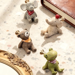 Könnte beinhalten: Eine Sammlung skurriler Tierknöpfe. Das Set enthält eine graue Maus, einen beigen Elefanten, einen braunen Bären und einen grünen Frosch, jeweils mit einem dekorativen goldfarbenen Knopf. Maus, Bär und Frosch tragen Bandanas.