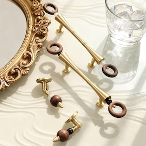 May include: Gold-toned cabinet hardware with wooden accents. The image shows several pieces, including long pulls and smaller knobs. The pulls have a cylindrical design with a wooden ring at each end. The knobs feature a wooden sphere and a decorative gold base.