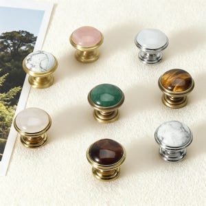 May include: A collection of decorative cabinet knobs with various stone and marble tops. The knobs feature brass or silver bases and come in colors like pink, white, green, brown, and tiger's eye.