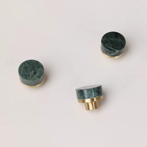 Może przedstawiać: Trzy zielone marmurowe gałki do szafek z mosiężnymi podstawami na białym tle. Gałki są cylindryczne, z polerowaną zieloną marmurową g&oacute;rą i złotą mosiężną podstawą. Gałki te są dekoracyjnym elementem okuć.