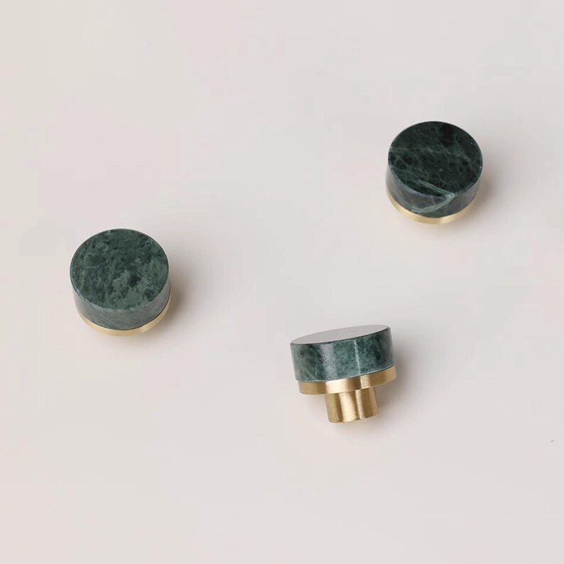 Może przedstawiać: Trzy zielone marmurowe gałki do szafek z mosiężnymi podstawami na białym tle. Gałki są cylindryczne, z polerowaną zieloną marmurową g&oacute;rą i złotą mosiężną podstawą. Gałki te są dekoracyjnym elementem okuć.