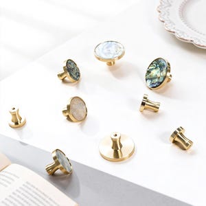 May include: A selection of cabinet knobs with gold-coloured bases and iridescent shell inlays. The knobs are in various shapes, including round and oval, and are arranged on a white surface. Some knobs are detached.