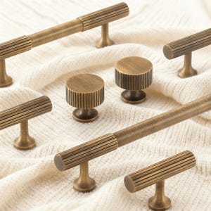 Può includere: Una collezione di ferramenta per mobili in ottone antico, tra cui maniglie a barra e pomelli rotondi. La ferramenta presenta un design a coste e una finitura calda e invecchiata. Le maniglie e i pomelli sono esposti su una superficie morbida e bianca.