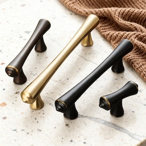 May include: A collection of cabinet hardware in various finishes. The selection includes bronze, gold, and black handles and knobs. The handles have a sleek, elongated design, while the knobs are shorter. All pieces feature a decorative ring detail.