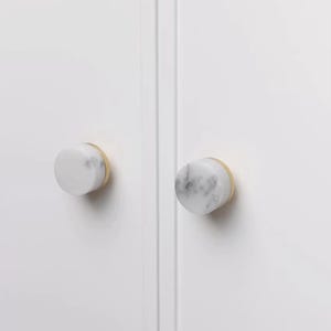 Może przedstawiać: Dwa uchwyty do szafek z białymi marmurowymi powierzchniami i podstawami w kolorze złotym są zamontowane na białych drzwiach szafki. Gałki są okrągłe i mają gładkie, polerowane wykończenie. Marmur ma szare żyłki.