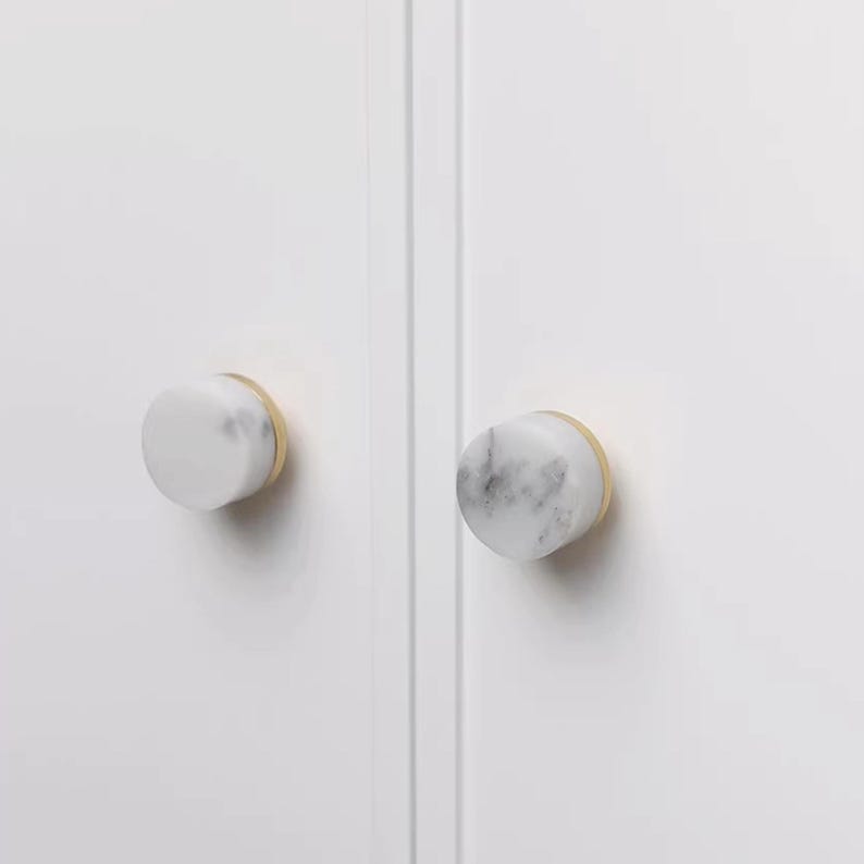 Może przedstawiać: Dwa uchwyty do szafek z białymi marmurowymi powierzchniami i podstawami w kolorze złotym są zamontowane na białych drzwiach szafki. Gałki są okrągłe i mają gładkie, polerowane wykończenie. Marmur ma szare żyłki.
