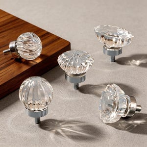May include: Five clear glass cabinet knobs with silver-colored bases. The knobs have various decorative ribbed and floral designs. The knobs are arranged on a light gray surface with a wooden cutting board in the background.