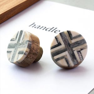 May include: Two decorative cabinet knobs with a geometric design. The knobs feature a wooden base and a circular top with a cross-like pattern in shades of gray, white, and cream. The word "handle" is printed on the paper.