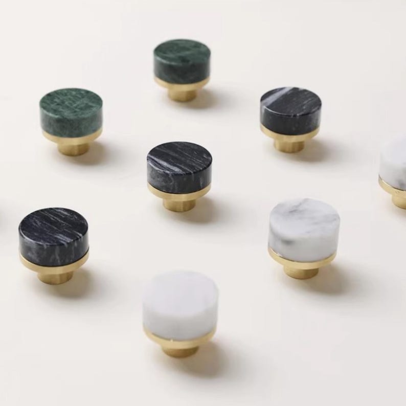 Może przedstawiać: Kolekcja marmurowych gałek do szafek w r&oacute;żnych kolorach, w tym zielonym, czarnym i białym. Każda gałka ma cylindryczną marmurową g&oacute;rę i złotą podstawę. Gałki są ułożone na białej powierzchni.