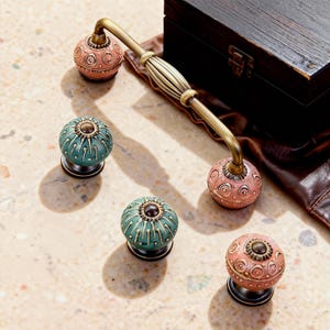 Op de afbeelding: Decoratieve kastbeslagset met een messing handgreep en vier keramische knoppen. De knoppen zijn roze en blauwgroen, met ingewikkelde patronen. De handgreep heeft een geribbeld middengedeelte. Voor meubels.