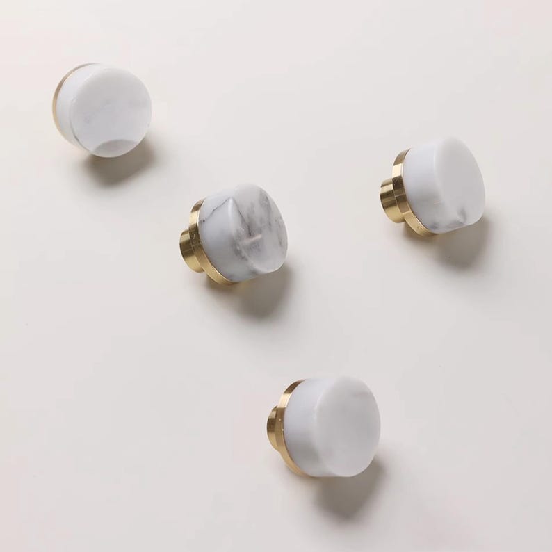 Może przedstawiać: Cztery białe marmurowe gałki do szafek ze złotymi podstawami ułożone są na białej powierzchni. Gałki mają cylindryczny kształt z płaskim wierzchem i złotą podstawą. Marmur ma szare żyłki.