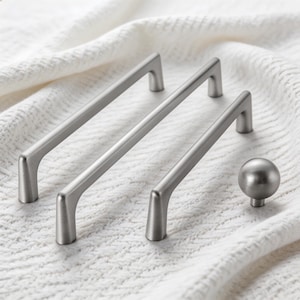 Könnte beinhalten: Drei Schrankgriffe aus gebürstetem Nickel und ein passender Knauf werden auf einer weißen, strukturierten Oberfläche präsentiert. Die Griffe haben ein schlankes, modernes Design mit einem geraden Stab und gebogenen Enden. Der Knauf hat eine einfache, runde Form.