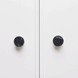 Może przedstawiać: Dwa okrągłe uchwyty do szafek z czarno-szarym wzorem marmuru zamontowane na białych drzwiach szafki. Uchwyty są wyśrodkowane na panelach drzwi, zapewniając minimalistyczną i nowoczesną estetykę.