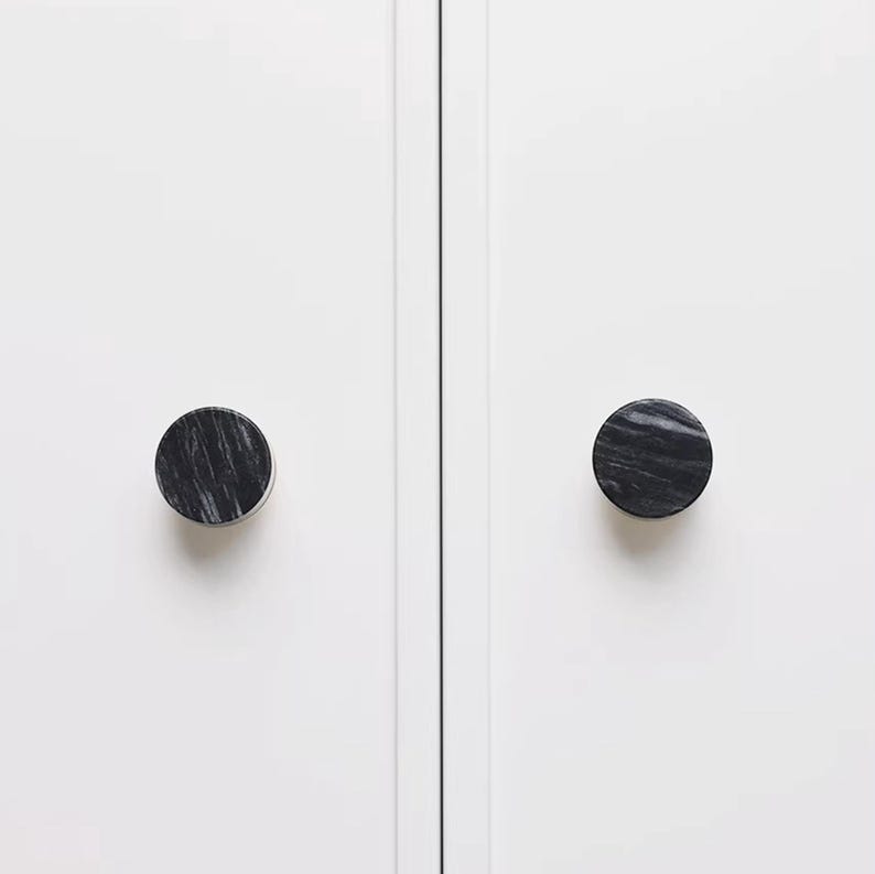 Może przedstawiać: Dwa okrągłe uchwyty do szafek z czarno-szarym wzorem marmuru zamontowane na białych drzwiach szafki. Uchwyty są wyśrodkowane na panelach drzwi, zapewniając minimalistyczną i nowoczesną estetykę.