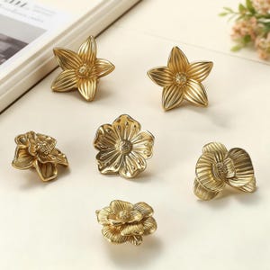 May include: Six gold-colored flower-shaped cabinet knobs. The knobs have detailed petal designs and varying shapes, including star-like and multi-layered floral forms. The hardware is arranged on a light-colored surface.