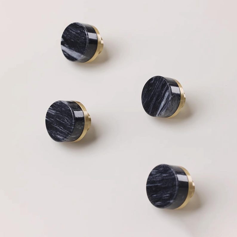 Może przedstawiać: Cztery dekoracyjne gałki do szafek. Każda gałka ma czarno-szarą marmurową powierzchnię ze złotym metalowym obramowaniem. Gałki są okrągłe i mają prosty, nowoczesny design. Są umieszczone na neutralnym tle.