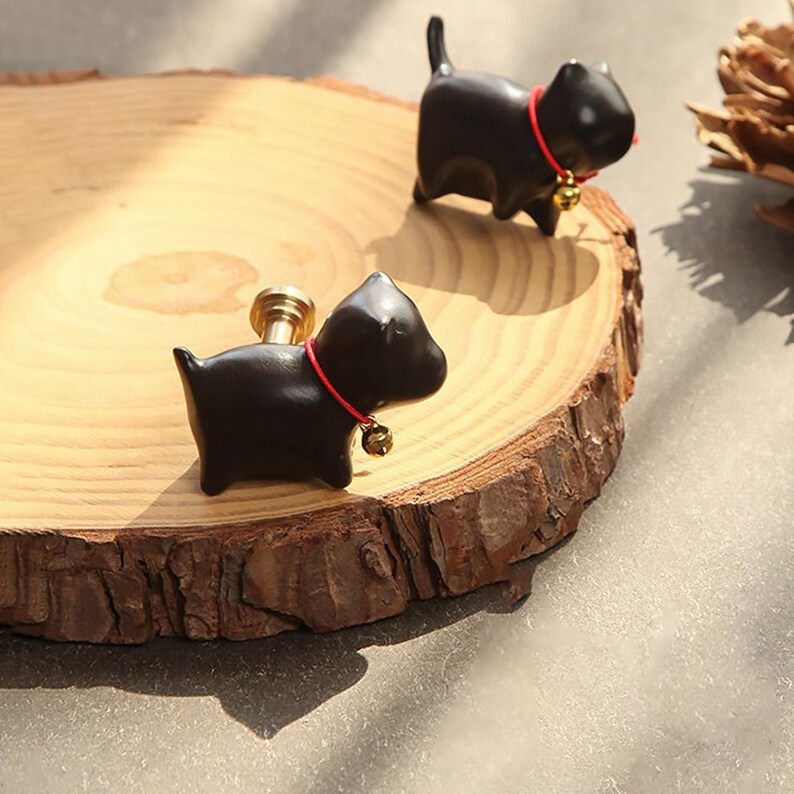 Puede incluir: Dos tiradores de armario negros con forma de gato, con collares rojos y peque&ntilde;as campanas doradas. Los tiradores est&aacute;n sobre una superficie de madera circular de color marr&oacute;n claro con un borde natural. Los tiradores tienen un v&aacute;stago dorado.