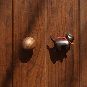 Puede incluir: Puerta de armario de madera con dos tiradores. Uno es un pomo redondo de madera, el otro es un tirador en forma de gato marr&oacute;n oscuro con un collar rojo y una peque&ntilde;a campana dorada. La puerta del armario es de madera oscura.