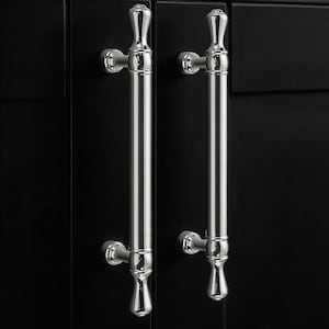 Könnte beinhalten: Zwei polierte Chrom-Schranktürgriffe. Jeder Griff hat einen langen, zylindrischen Griff mit dekorativen Details oben und unten. Die Griffe sind an einer schwarzen Schranktür montiert.