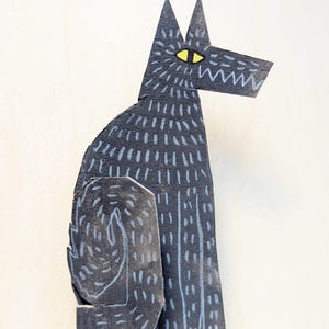 May include: A black paper cutout of a wolf with a yellow eye and white accents. The wolf is sitting with its head turned to the right. The paper has a textured appearance with white dashes.