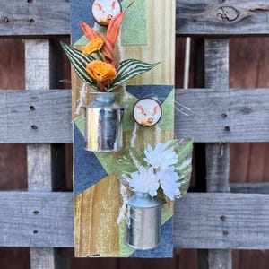 May include: A rustic wooden wall hanging featuring two small metal milk cans filled with faux flowers. The wooden board has denim and patterned fabric accents, along with two pig illustrations. The flowers are orange and white.