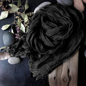 May include: A black scarf with a fringed edge is artfully arranged on a dark surface. The scarf is draped over a piece of wood, with small stones and sprigs of greenery and pink flowers adding a natural touch to the composition.