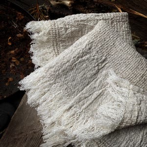May include: A folded, off-white woven throw blanket with a diamond pattern and fringed edges. The blanket is made of a soft, textured material and is draped over a wooden surface. The fringe is a bright white color.