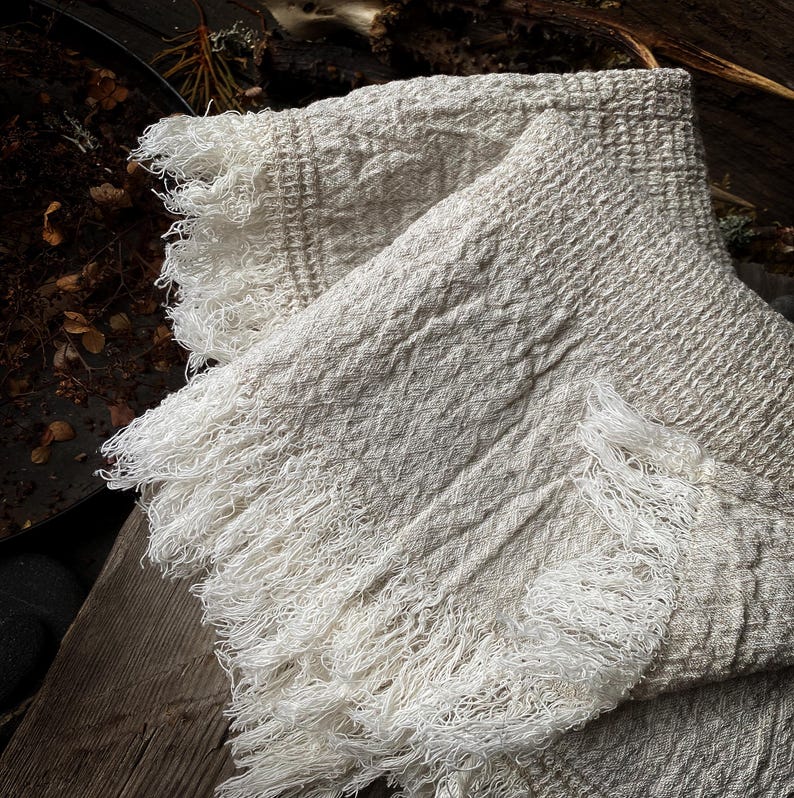 May include: A folded, off-white woven throw blanket with a diamond pattern and fringed edges. The blanket is made of a soft, textured material and is draped over a wooden surface. The fringe is a bright white color.