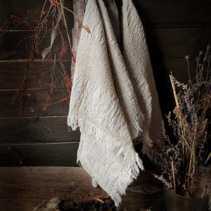 May include: Off-white, textured linen towels with fringed edges hang against a dark wood background. Dried branches and plants are arranged nearby, creating a rustic, natural aesthetic. The towels appear soft and slightly wrinkled.
