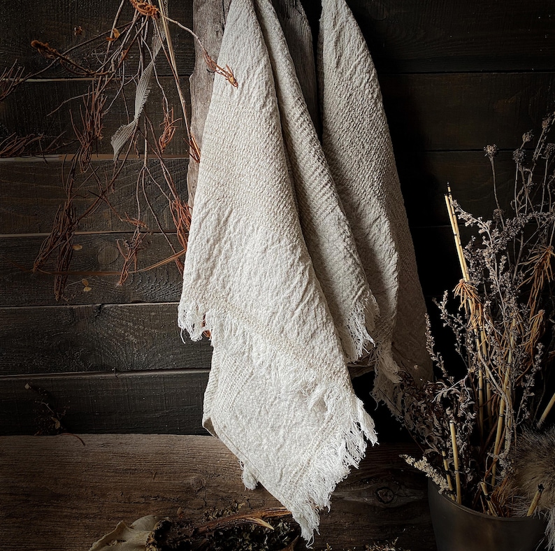 May include: Off-white, textured linen towels with fringed edges hang against a dark wood background. Dried branches and plants are arranged nearby, creating a rustic, natural aesthetic. The towels appear soft and slightly wrinkled.