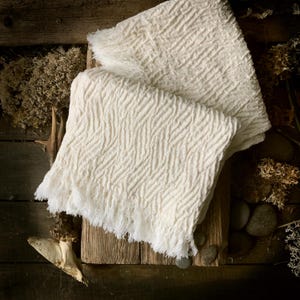 Puede incluir: Manta de color crema con textura y borde con flecos. La manta tiene un patrón tejido y está doblada sobre una superficie de madera. La manta está hecha de un material suave y es adecuada para la decoración del hogar.