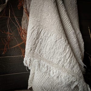 May include: A cream-colored, textured blanket with a fringed edge. The blanket is draped, revealing its woven pattern and soft texture. The background features dark wood and dried branches, enhancing the rustic aesthetic.