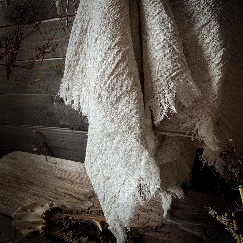 May include: Off-white, textured throw blanket with fringed edges. The blanket is draped over a wooden surface, showcasing its woven pattern and soft texture. The neutral color and rustic aesthetic suggest a cozy home decor item.