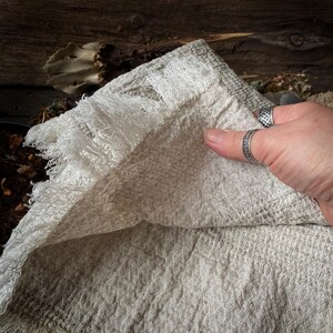 May include: A close-up of a beige linen towel with a diamond pattern and fringed edges. The towel is folded, revealing its texture and construction. The neutral color and natural fibers suggest a rustic or farmhouse style.