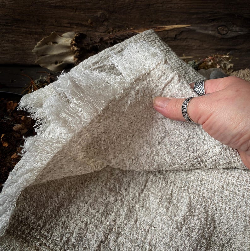 May include: A close-up of a beige linen towel with a diamond pattern and fringed edges. The towel is folded, revealing its texture and construction. The neutral color and natural fibers suggest a rustic or farmhouse style.