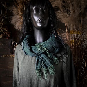 Può includere: Sciarpa verde scuro con frange drappeggiata attorno al collo di un manichino. La sciarpa è realizzata in un tessuto strutturato ed è abbinata a una camicia verde oliva a maniche lunghe. Il manichino ha i capelli neri ed è su uno sfondo rustico.