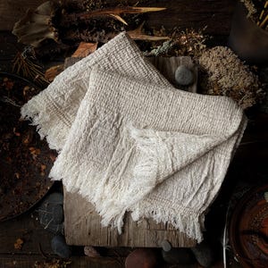 May include: Two off-white, textured linen towels with fringed edges are folded on a rustic wooden surface. The towels have a woven pattern and a soft, natural appearance. The background includes natural elements like wood, stones, and foliage.